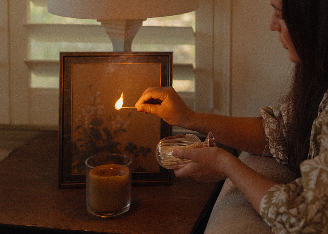 Woman lighting a candle next to a framed picture on a table with a lamp in the background.