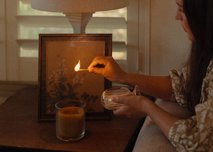 Woman lighting a candle next to a framed picture on a table with a lamp in the background.