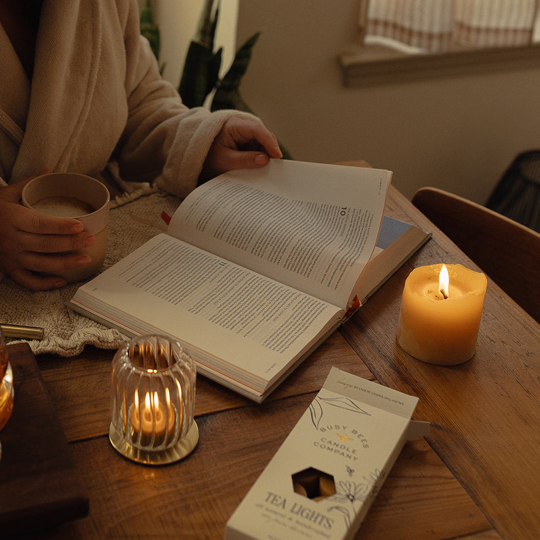 Person in a robe reading a book with a lit candle and tea set on a wooden table.