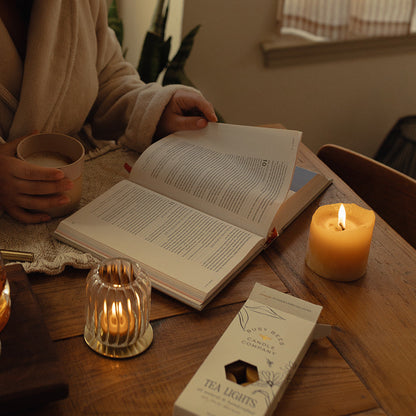 Person in a robe reading a book with a lit candle and tea set on a wooden table.
